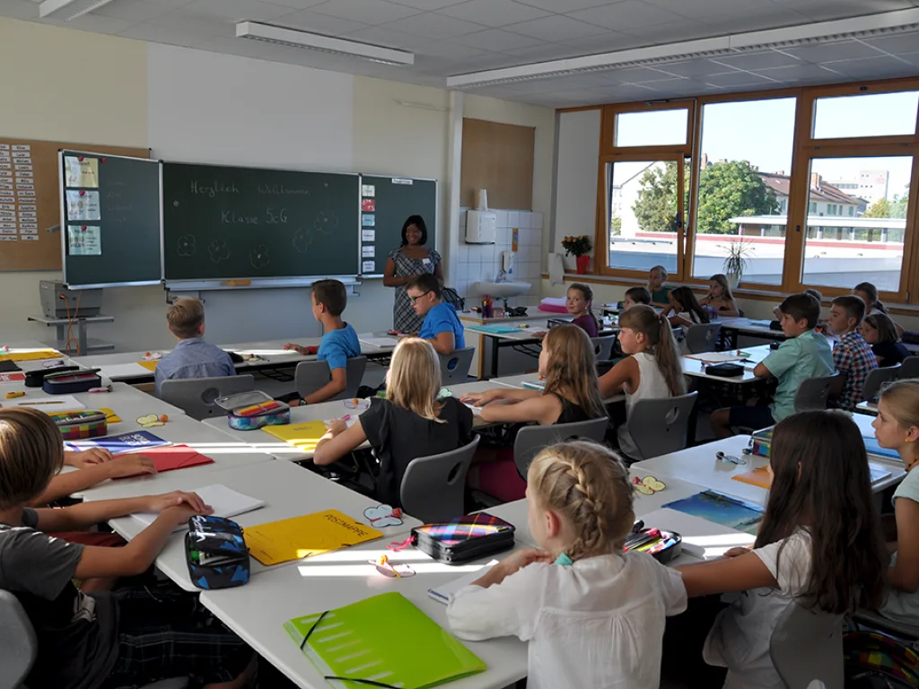 Eine Lehrkraft steht vorn, die Kinder sitzen an ihren Plätzen.